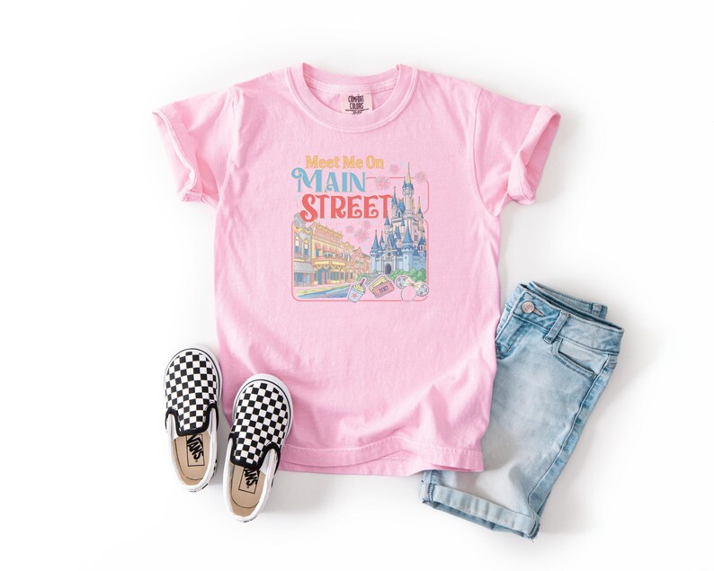 May include: A light pink t-shirt with the words "Meet Me On Main Street" and a colorful graphic of a castle and buildings. The shirt is paired with blue denim shorts and black and white checkered slip-on shoes.
