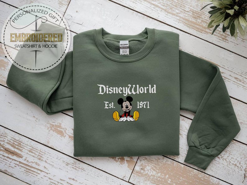 May include: An olive green sweatshirt featuring the embroidered text "DisneyWorld" and "Est. 1971" in white. A cartoon Mickey Mouse image is embroidered below the text. The sweatshirt is folded on a light wood surface.