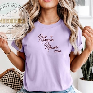 May include: A light purple t-shirt with the words "Minnie Mouse xoxo" in a cursive font. The shirt also has a circular graphic that says "Personalized Gift" and "Embroidered Comfort Colors".