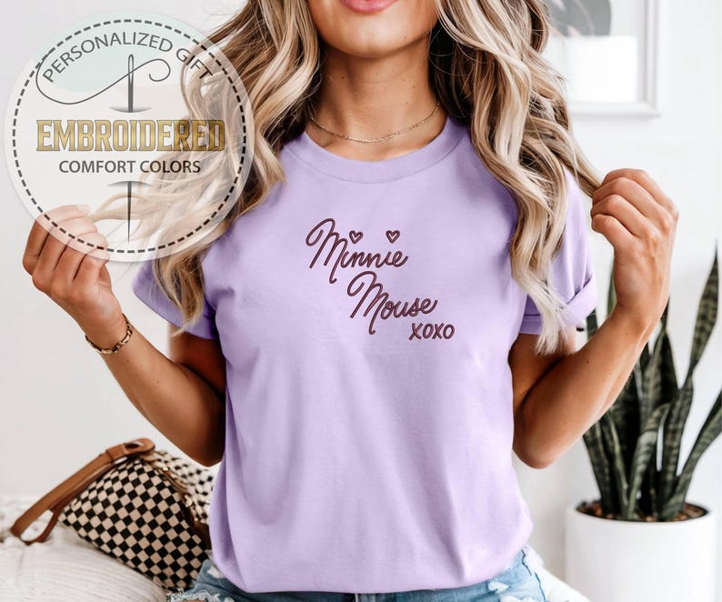 May include: A light purple t-shirt with the words "Minnie Mouse xoxo" in a cursive font. The shirt also has a circular graphic that says "Personalized Gift" and "Embroidered Comfort Colors".