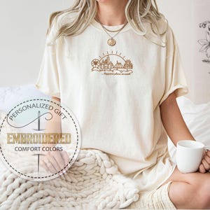 May include: Cream-colored t-shirt with a brown embroidered design of a castle, Ferris wheel, and sun, with the text "happiest place on earth." The shirt also features a circular logo that says "Personalized Gift" and "Embroidered Comfort Colors."