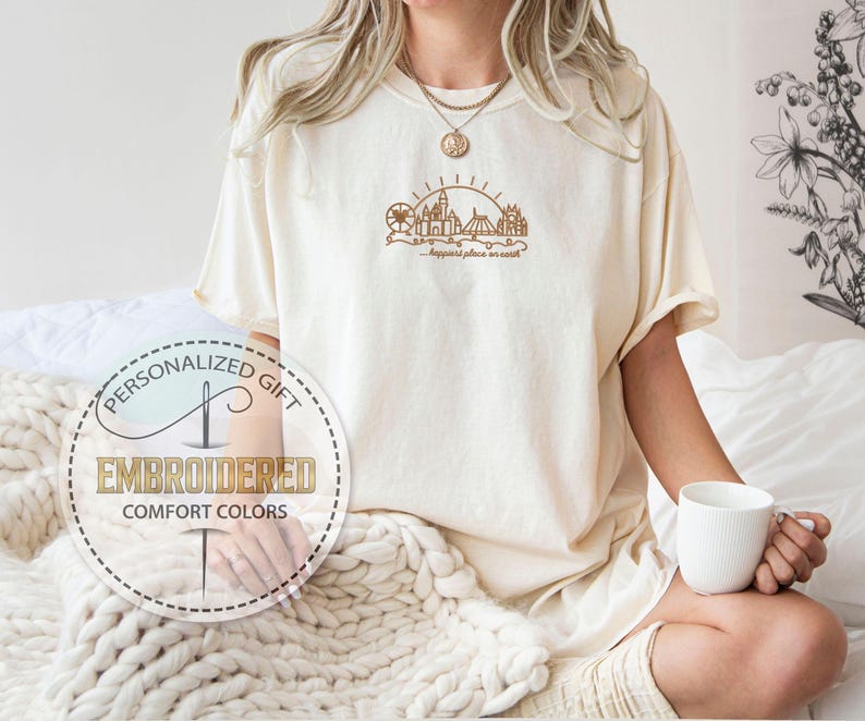 May include: Cream-colored t-shirt with a brown embroidered design of a castle, Ferris wheel, and sun, with the text "happiest place on earth." The shirt also features a circular logo that says "Personalized Gift" and "Embroidered Comfort Colors."