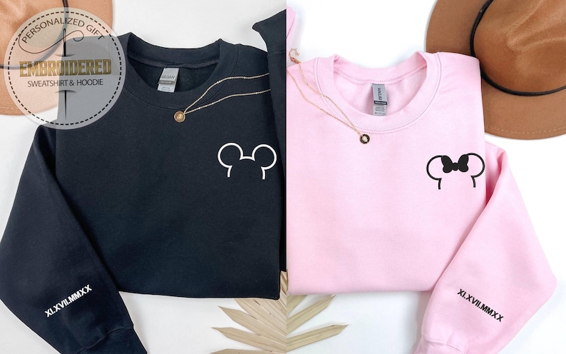 以下が含まれることがあります： 白のミッキーマウスとミニーマウスの耳のデザインが施された黒とピンクのスウェットシャツ。各スウェットシャツには金のネックレスと、袖にはローマ数字のXLXVIIIMMXXが付いています。左上には「Personalized Gift EMBROIDERED SWEATSHIRT & HOODIE」と書かれた丸いロゴがあります。