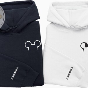 以下が含まれることがあります： 紺色と白の2つのフード付きスウェットシャツ。紺色のスウェットシャツには白いミッキーマウスの耳のデザインがあり、白いスウェットシャツにはミニーマウスのリボンのデザインがあります。どちらも袖に「XL.XVII.MMXX」の文字が入っています。画像には「PERSONALIZED GIFT EMBROIDERED SWEATSHIRT & HOODIE」の文字も含まれています。