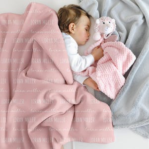 May include: A soft pink blanket featuring the repeated white text LORAN MILLER in a script font, paired with a light pink plush lion security blanket with a textured waffle-knit fabric, resting alongside a soft grey plush blanket.