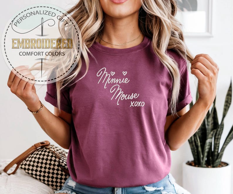 May include: A maroon t-shirt with the words "Minnie Mouse" in white script, with two small hearts and "XOXO" underneath. The shirt has rolled-up sleeves and is part of a personalized gift collection.