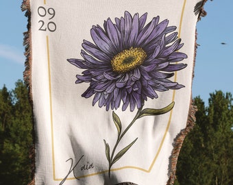 May include: A woven tapestry with a purple flower and green stem. The tapestry has a white background with a gold frame and the text "09 20" and "Kaia" in black.