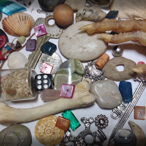 Könnte beinhalten: Eine Sammlung verschiedener Objekte, darunter Muscheln, Würfel, Schlüssel, Steine, Perlen, ein Knochen und eine kleine Glasflasche mit getrocknetem Material. Ein Vogelfuß ist ebenfalls vorhanden. Die Gegenstände sind auf einer weißen Oberfläche verstreut.