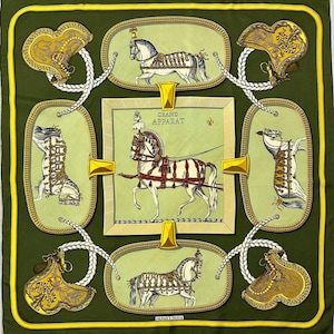 Puede incluir: Un pañuelo cuadrado de seda en tonos verde oliva, dorado y blanco. El pañuelo presenta ilustraciones enmarcadas de caballos y sillas de montar, con el texto "GRAND APPARAT" y "HERMES - PARIS". El borde es amarillo contrastante.