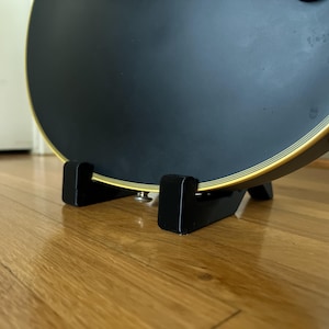 以下が含まれることがあります： 黒いボディと金色のトリムが付いたアコースティックギター。黒い木製のスタンドに置かれています。ギターのサウンドホールは見えません。スタンドは木の床にあります。