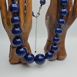 May include: A navy blue beaded necklace featuring large, round beads. The necklace has a silver-toned clasp and an adjustable chain. The beads are evenly spaced and have a glossy finish. The necklace is displayed on a wooden hand.