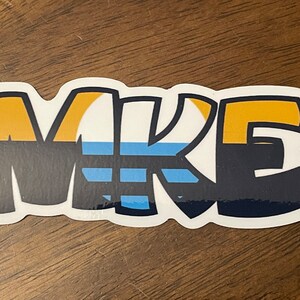 May include: A white sticker with the word "MIKE" in black, outlined in dark blue. The letters are stylized with a yellow and blue stripe pattern.