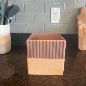 May include: A square-shaped container featuring a two-tone design. The upper portion is a mauve-colored section with vertical ribbed detailing, while the lower portion is a solid, textured light peach base with a matte finish.
