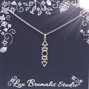 Puede incluir: Un collar con colgante plateado con un diseño geométrico, que incluye triángulos, un círculo y líneas horizontales. El collar cuelga de una fina cadena sobre un fondo azul oscuro con patrones florales y de estrellas blancas. El texto "Lux Brumalis Studio" está en la parte inferior.