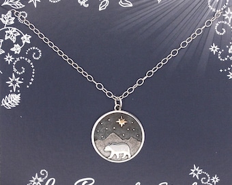 Pode incluir: Um colar de prata com um pingente redondo que apresenta uma silhueta de urso estilizada sobre um fundo azul escuro com uma estrela dourada. O pingente é rodeado por uma corrente de prata.