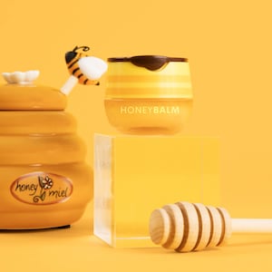 May include: A yellow and brown honey pot with a bee on the lid and the words "honey miel" on the side. Next to it is a yellow and brown honey balm jar with the words "HONEYBALM" on the front. A wooden honey dipper is on a clear acrylic block.