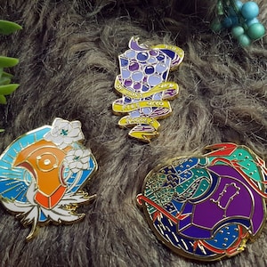 May include: Three enamel pins with gold accents. The first pin features a purple and blue design with a banner that reads "Fire, Water, Earth, Air". The second pin features a blue, orange, and white design with white flowers. The third pin features a purple, blue, green, and red design.