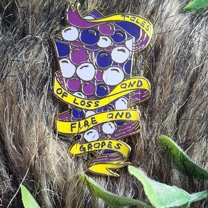 May include: A gold-colored enamel pin with a purple and blue grape design. The pin has a yellow banner that reads "Tales of Loss and Fire and Grapes".
