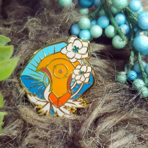 May include: A gold-toned enamel pin featuring a stylized bird with orange, red, and white accents. The bird is surrounded by blue and white enamel, and white flowers.