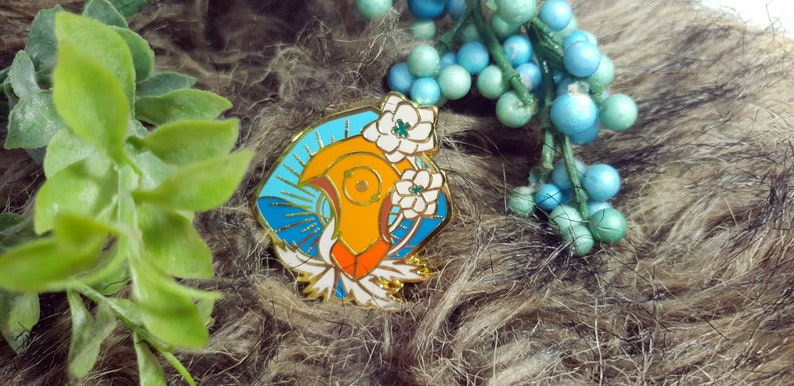 May include: A gold-toned enamel pin featuring a stylized bird with orange, red, and white accents. The bird is surrounded by blue and white enamel, and white flowers.