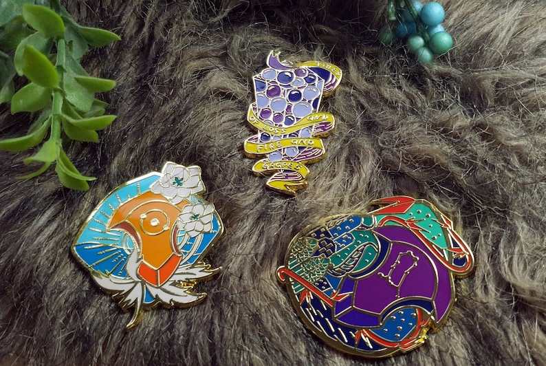May include: Three enamel pins with gold accents. The first pin features a purple and blue design with a banner that reads "Fire, Water, Earth, Air". The second pin features a blue, orange, and white design with white flowers. The third pin features a purple, blue, green, and red design.