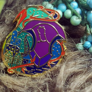 May include: A gold-toned enamel pin with a colorful abstract design. The pin features a purple, blue, and green design with red accents.