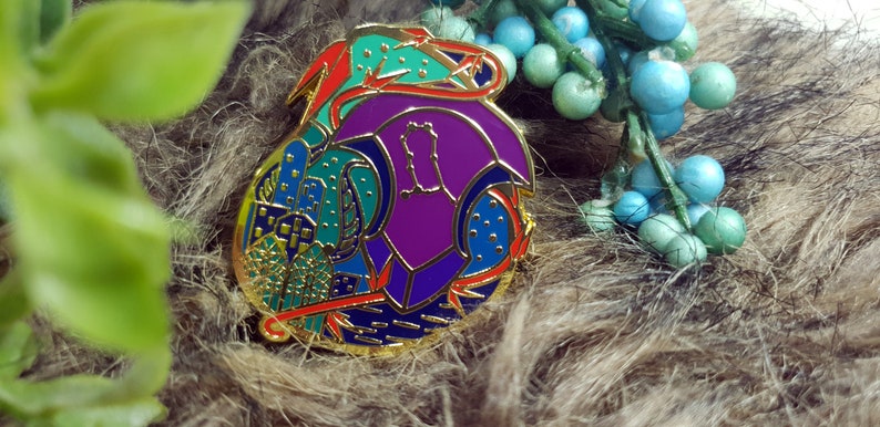 May include: A gold-toned enamel pin with a colorful abstract design. The pin features a purple, blue, and green design with red accents.