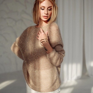 Puede incluir: Una mujer con un suéter beige brillante de manga larga y pantalones blancos. Está de pie frente a una pared blanca con una cortina blanca detrás de ella.