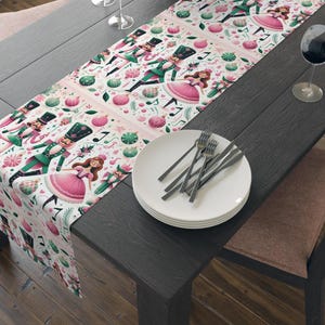May include: A white table runner with a pink and green Christmas theme. The runner features a repeating pattern of nutcracker soldiers, ballerinas, and musical notes.