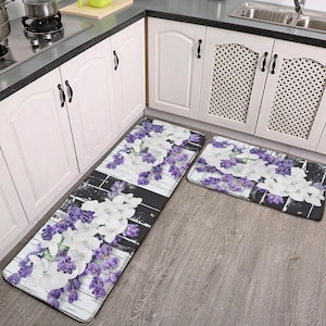 Puede incluir: Un juego de dos alfombras de cocina con un diseño floral. Las alfombras tienen un fondo a rayas blanco y negro con flores moradas y blancas. Las alfombras se colocan delante de un armario de cocina blanco.