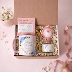 Op de afbeelding: Een geschenkdoos met een witte keramische mok met blauwe bloemaccenten en de tekst Angie's Bridal Shower, een roze Tea With Tae theezakje, een goudkleurige lepel, een ronde roze badbom en een klein glazen potje met een kaars en label.