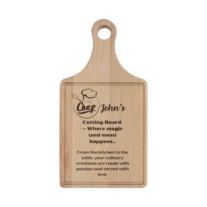 May include: A light-coloured wooden cutting board with a handle and a circular hole. It features a laser-engraved chef hat icon, the text "Chef John's Cutting Board – Where magic (and mess) happens.." and a message about culinary creations.