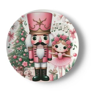 May include: A white ceramic plate featuring a pink and green Christmas design. A pink nutcracker with a gold hat and a pink ballerina with a flower crown are depicted on the plate.