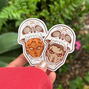 May include: Two stickers featuring skull designs with faces inside. One sticker has the text "one end" and the other has "one flesh". The faces are detailed with different skin tones and hair colors. The stickers are held in front of a blurred green and pink background.