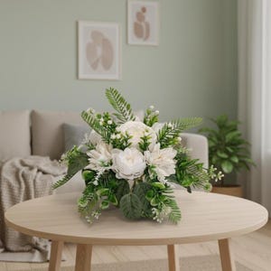 May include: A floral arrangement featuring white peonies and dahlias, accented with green leaves and sprigs. The bouquet is displayed on a light wood table, with a neutral-toned background. The arrangement is a centerpiece.