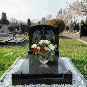 May include: A black granite headstone with an engraved cross and angel figures. A white vase holds red and white roses. The headstone is in a cemetery with other headstones and green grass.