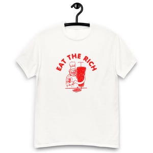 May include: White t-shirt with a red graphic. The graphic features a chef with a beard and chef's hat, holding a curved knife next to a vertical stack of meat. The text "EAT THE RICH" is above the chef.
