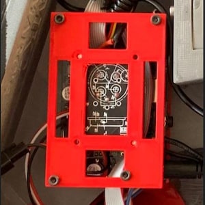 May include: A red rectangular frame with cutouts surrounds a black circuit board with intricate white designs. Wires and cables in various colors are visible, along with screws securing the frame. The image showcases electronic components.