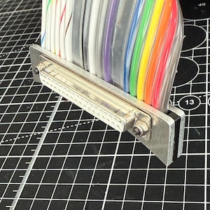May include: A multi-colored ribbon cable featuring individual wires in white, blue, purple, green, yellow, and orange, connected to a metallic D-subminiature connector with two visible mounting screws and a rectangular silver-toned metal bracket.