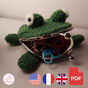 May include: Green crocheted frog coin purse with white eyes and a silver metal clasp. The purse is open and filled with colorful rubber bands.