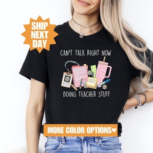 May include: Black t-shirt with a pink and white graphic of teacher supplies and the text "Can't talk right now doing teacher stuff".