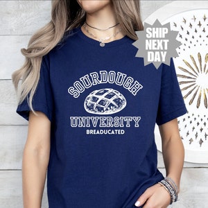 Può includere: Una maglietta blu navy con testo bianco che recita "Sourdough University" e "BREADUCATED". Sopra il testo è presente un'illustrazione bianca di una pagnotta di pane a lievitazione naturale.