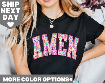 Puede incluir: Camiseta negra con la palabra "AMEN" en un diseño de estampado de leopardo colorido, contorneado en rosa. La camiseta tiene cuello redondo y mangas cortas. La imagen también incluye el texto "MORE COLOR OPTIONS".