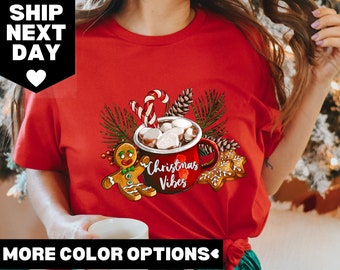 Op de afbeelding: Rood T-shirt met een kerstthema met een mok warme chocolademelk met marshmallows, een peperkoekman, snoepstokken en dennentakken. De tekst "Christmas Vibes" staat op de mok.