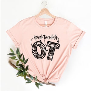 Può includere: Una maglietta rosa con il testo "spooktacular 07" in nero. I numeri sono progettati per assomigliare a ragnatele con piccoli ragni neri.