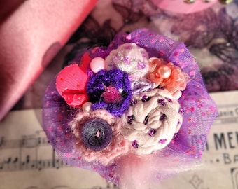 May include: A small bouquet of fabric flowers in shades of pink, purple, and white. The flowers are arranged in a tulle and lace base with a white feather accent.