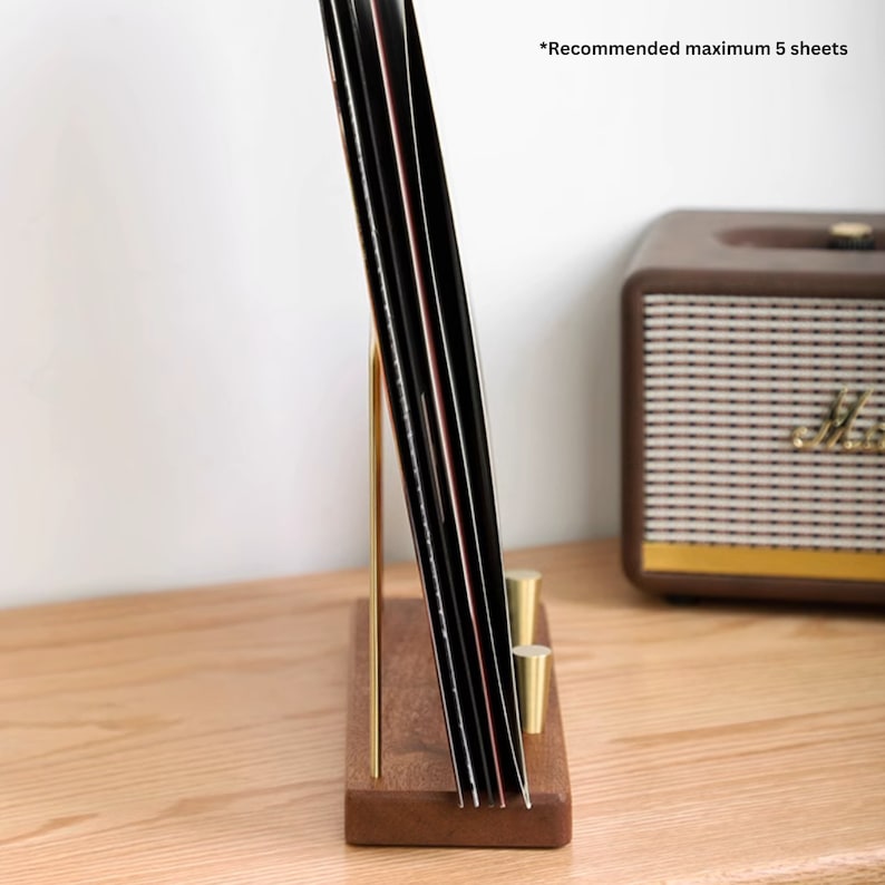 May include: A wooden and brass record holder with a gold-colored support. The holder is designed to display vinyl records. The text "Recommended maximum 5 sheets" is visible. A brown speaker is in the background.