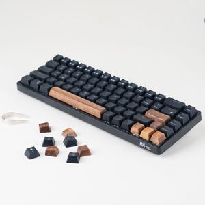 May include: A black mechanical keyboard with wooden detailing. The keyboard features black keycaps and a wooden space bar. Several wooden and black keycaps are scattered nearby. A pair of silver tweezers is also visible.