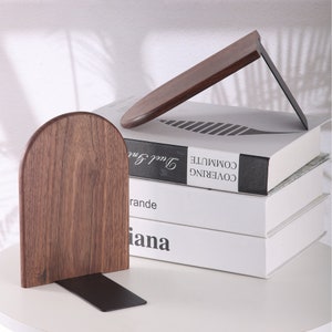 Pu&ograve; includere: Due fermalibri in legno con finitura marrone scuro e base in metallo nero. Uno &egrave; in posizione verticale, l'altro angolato, che sostiene una pila di libri. I libri hanno copertine bianche e testo nero.