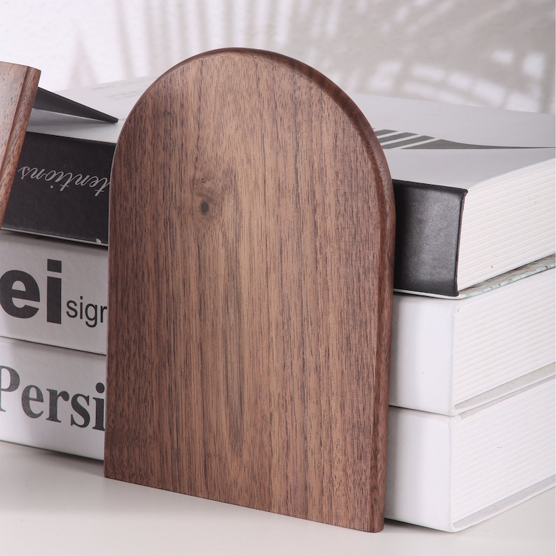 Pu&ograve; includere: Un paio di fermalibri in legno con finitura marrone scuro. I fermalibri hanno una parte superiore arrotondata e una base rettangolare. Sono posizionati accanto a una pila di libri bianchi.